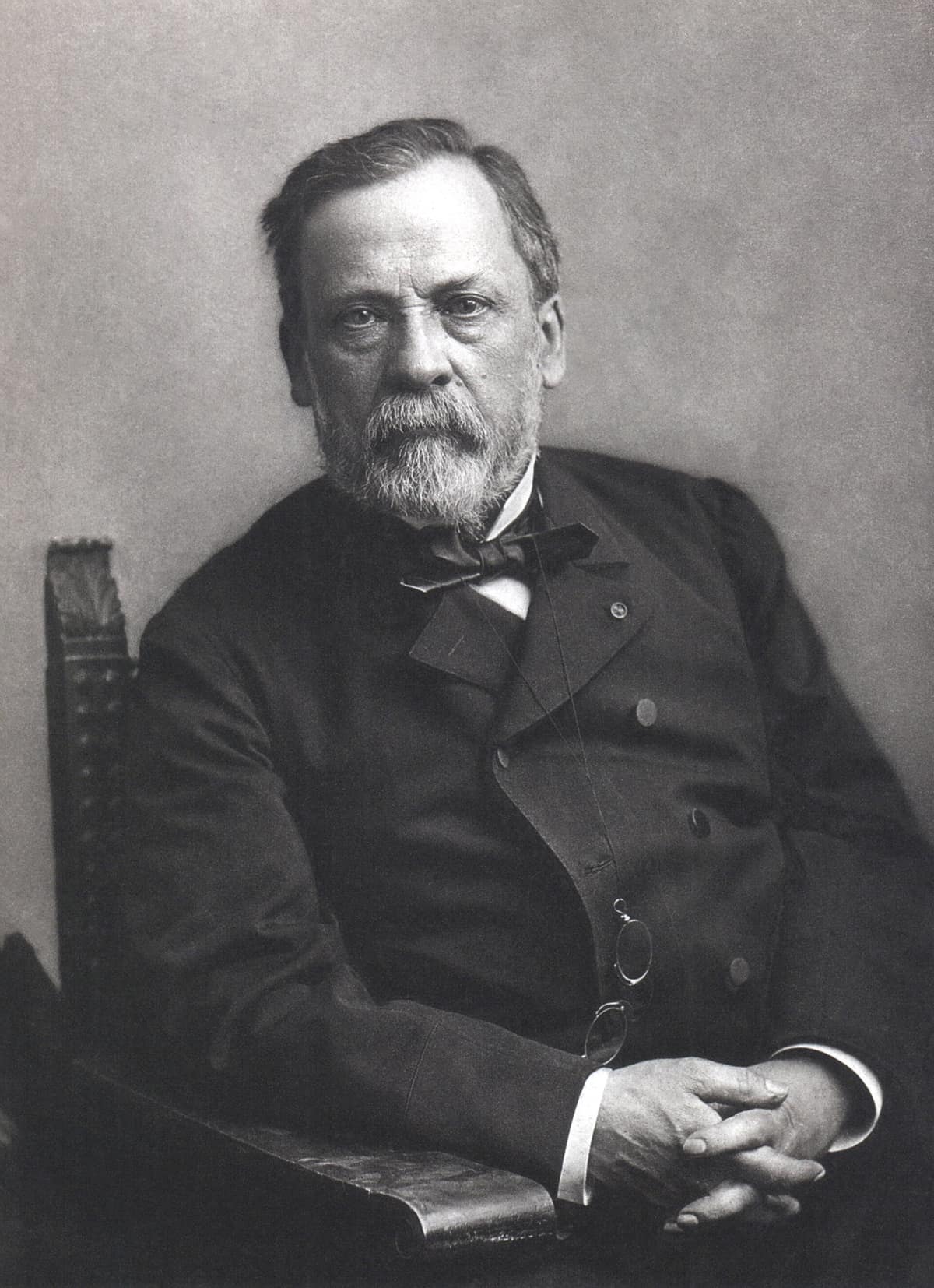 Black-and-white studio photo of a bearded old man in a dark suit and bow tie, seated with hands folded