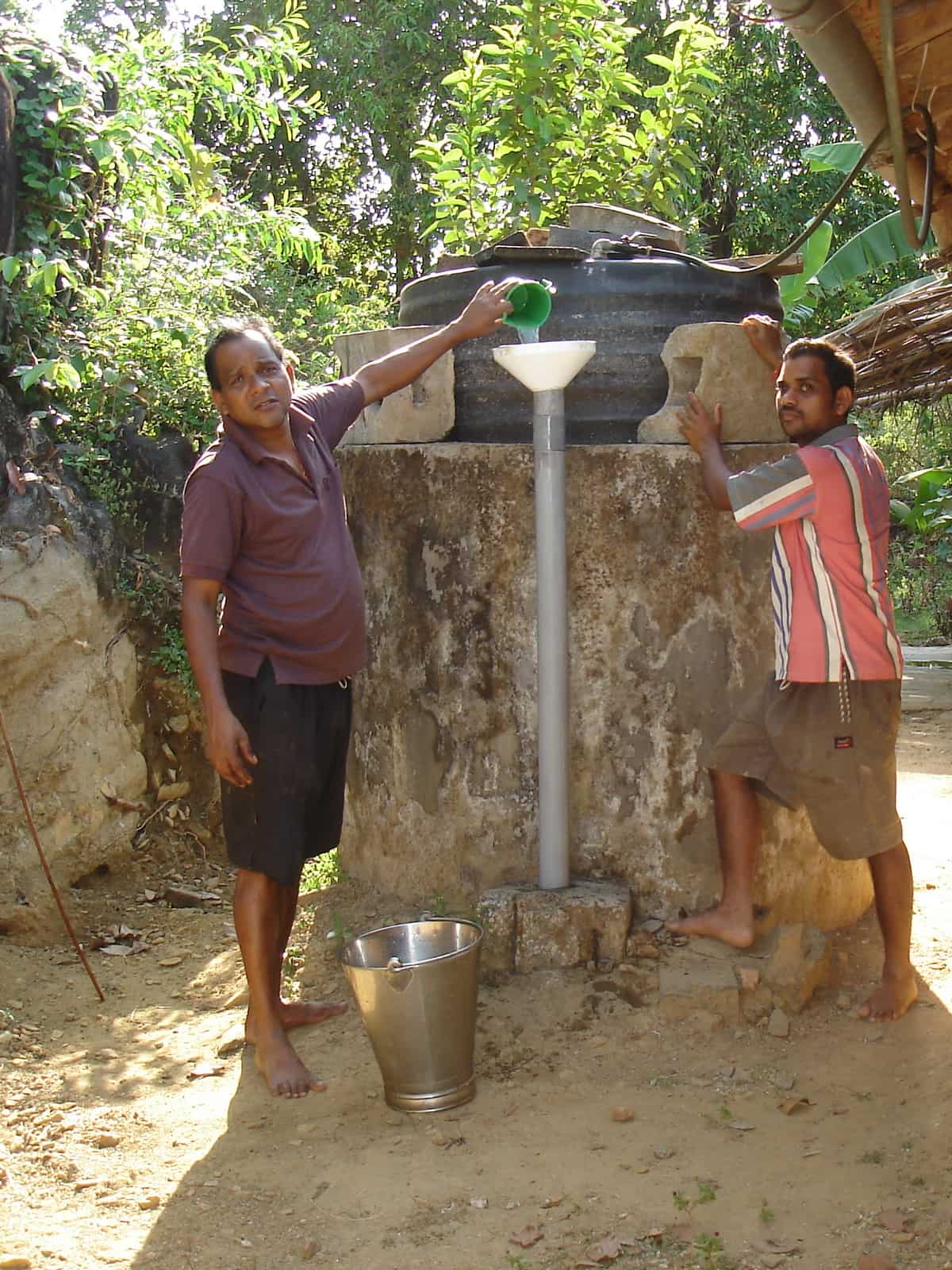 Two men at a small round household biogas plant; one pours slurry from a jug into a funnel and pipe that feed the tank