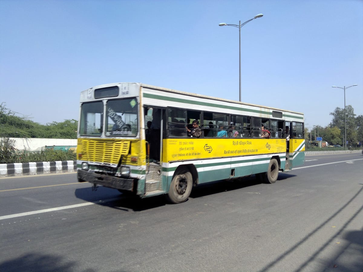 A yellow and green Delhi Transport Corporation bus with passengers, driving on a wide city road