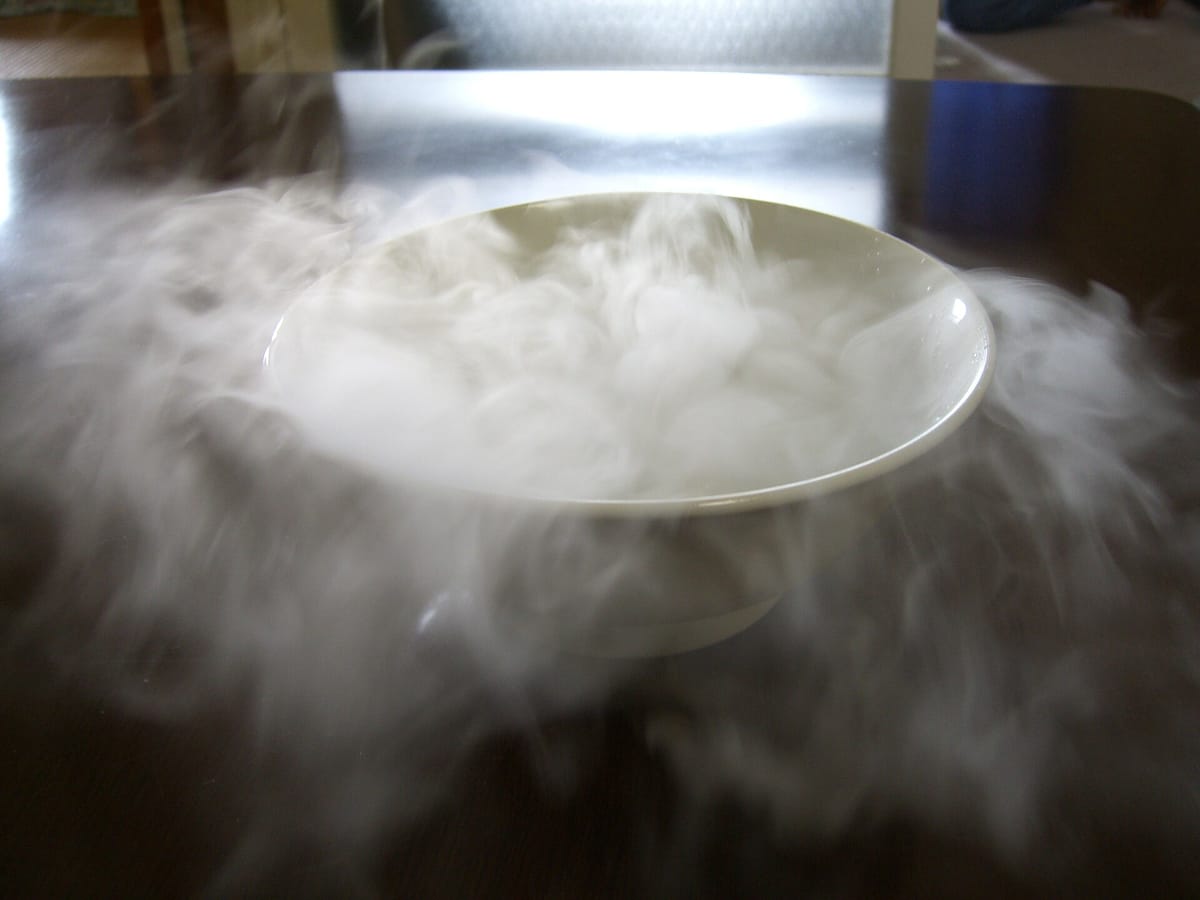 A white bowl of dry ice overflowing with thick white fog that spreads across a dark table