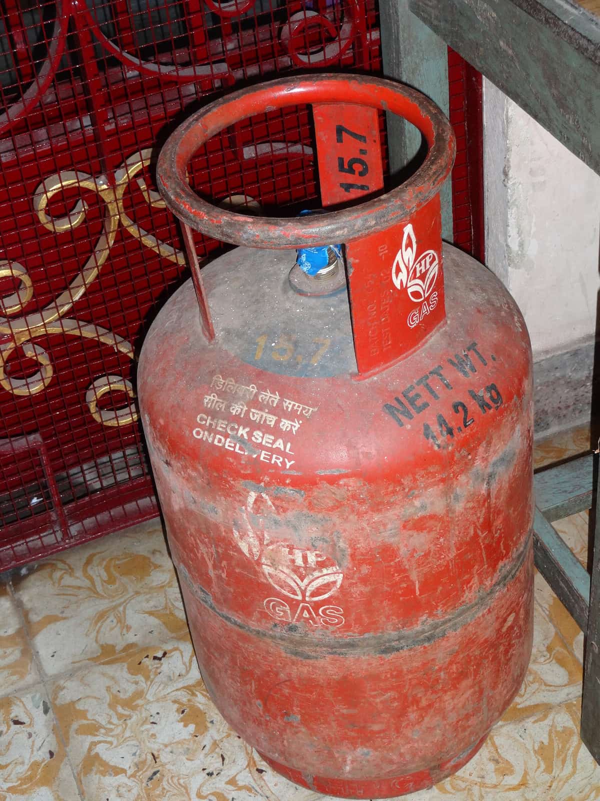 A red domestic LPG cylinder marked 'Nett Wt. 14.2 kg' with a handle ring and a sealed valve, standing on a tiled floor
