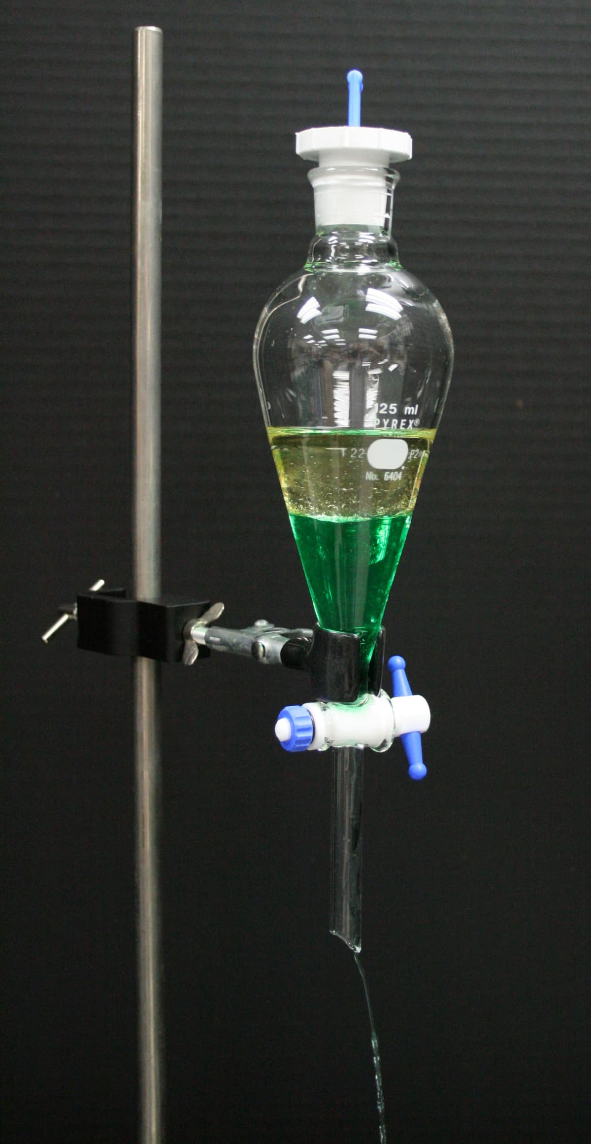 A glass separating funnel on a stand with a yellow oil layer floating above a green water layer
