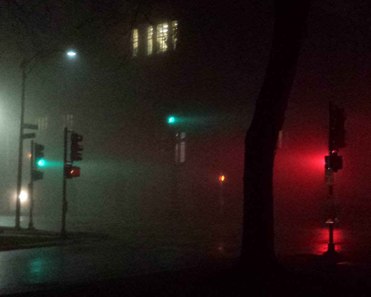 A street corner at night in thick fog; beams of green and red light spread out around the traffic signals