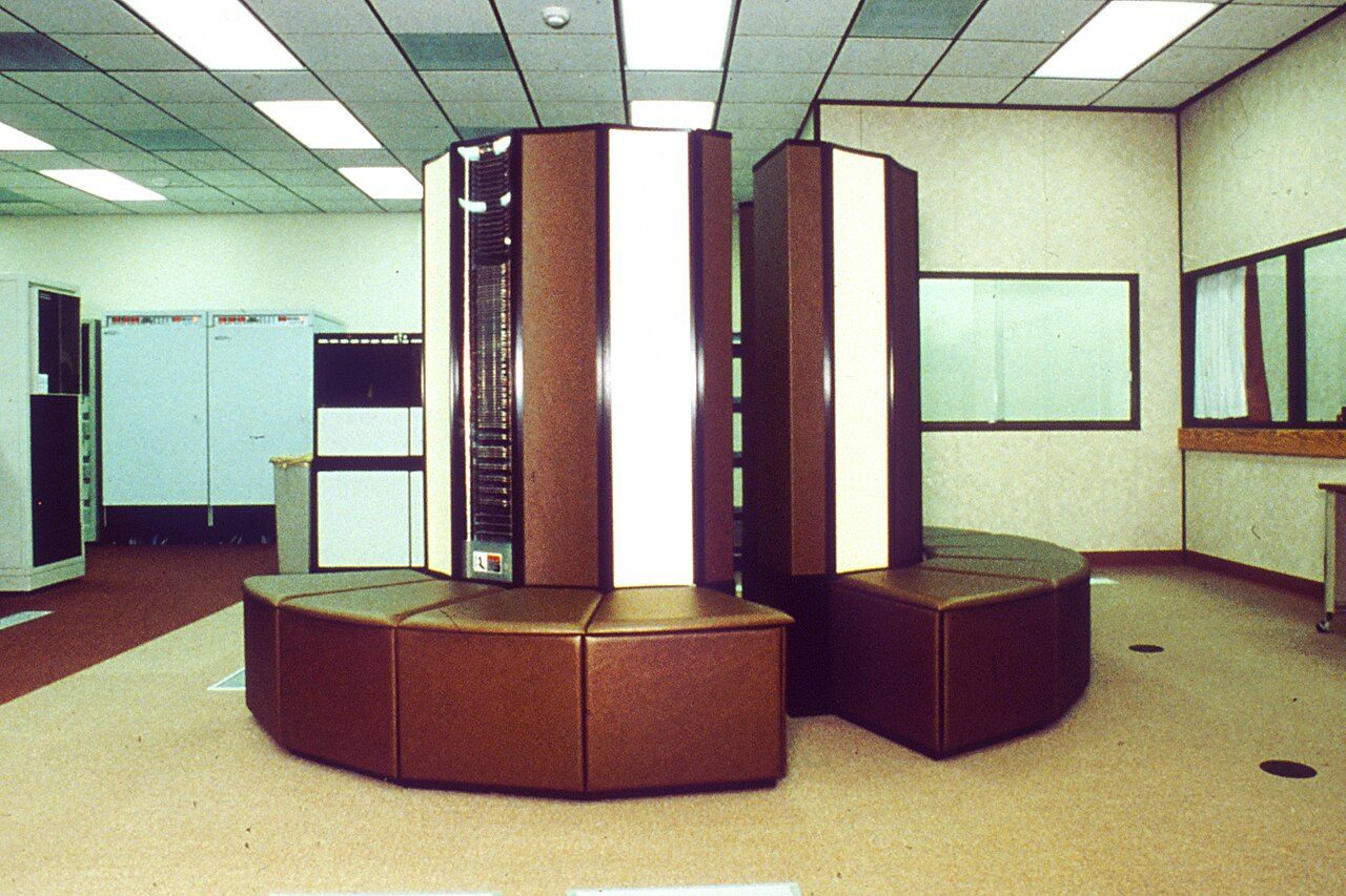 A tall round Cray supercomputer with a bench-like base in a computer room
