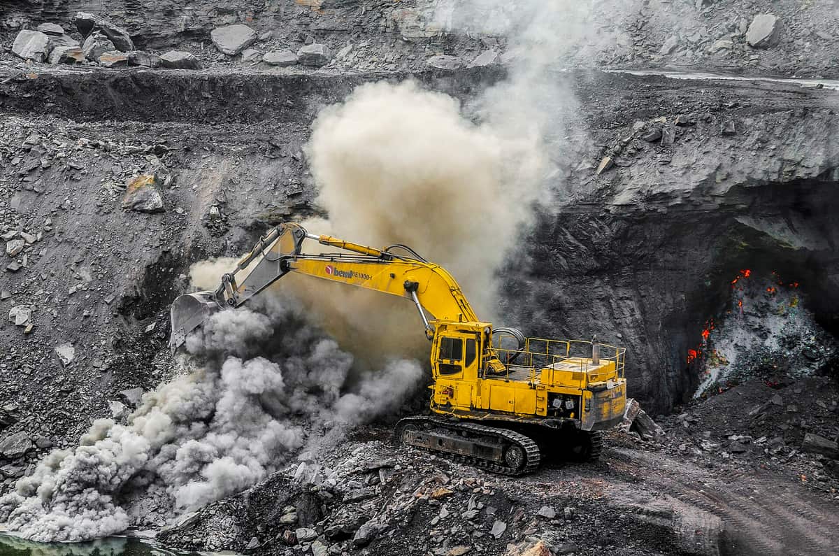 A large yellow excavator digging in a grey open-pit mine, with clouds of dust and a patch of glowing red fire in the rock wall