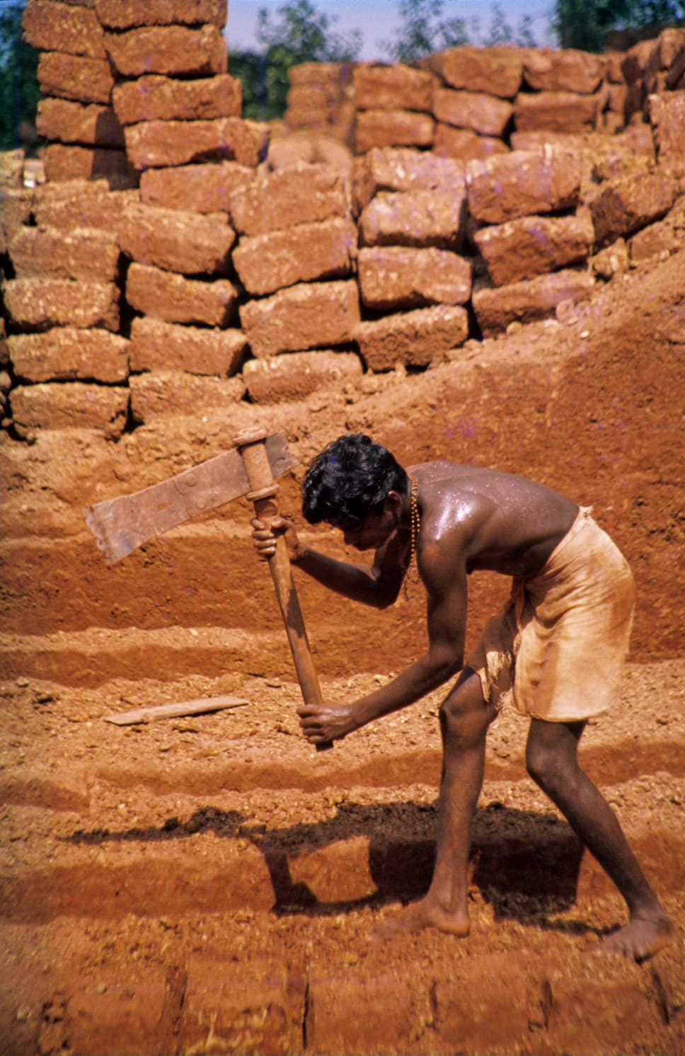 A worker swinging a heavy pick to cut blocks from red earth, with a wall of stacked red stone blocks behind him