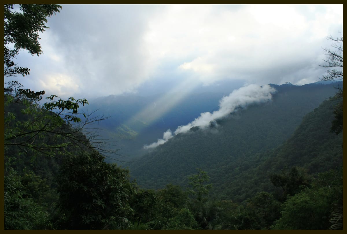 Dark rain clouds and a band of white mist over steep green forested hills, with a beam of sunlight breaking through