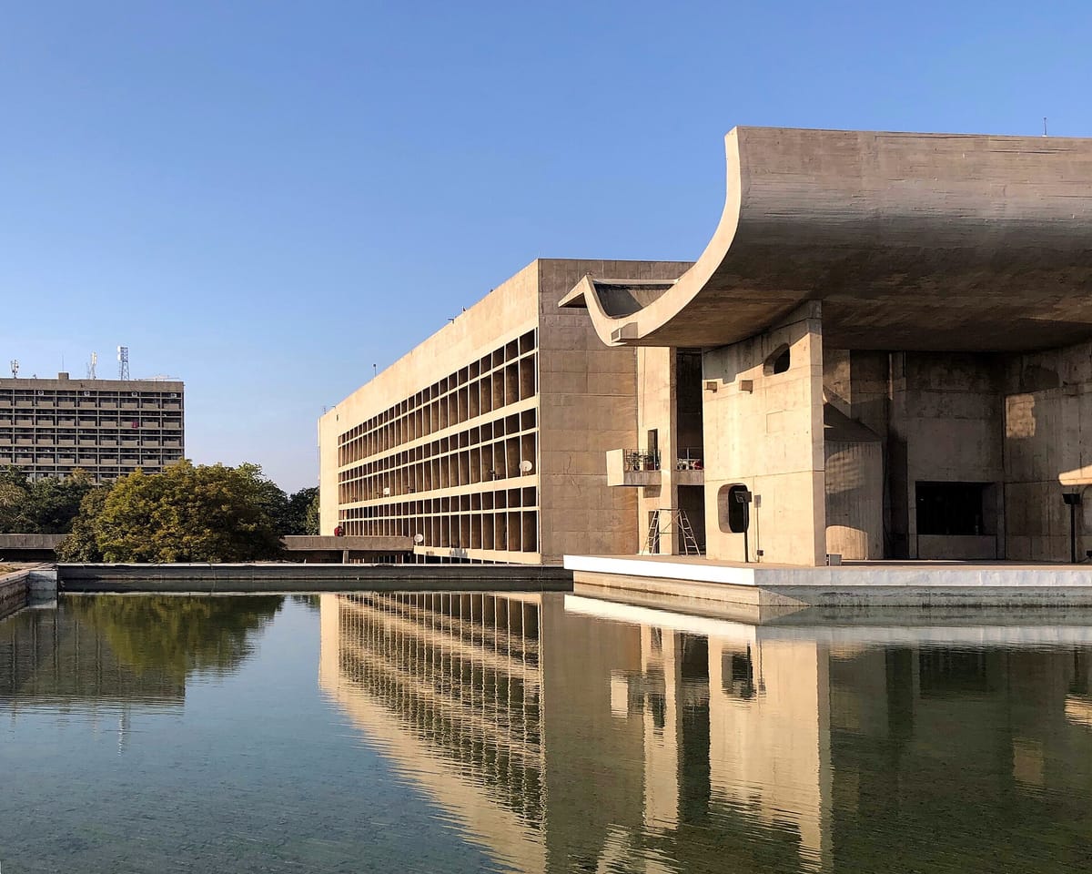A large bare-concrete building with a huge curved roof canopy, reflected in a still pool under a clear blue sky