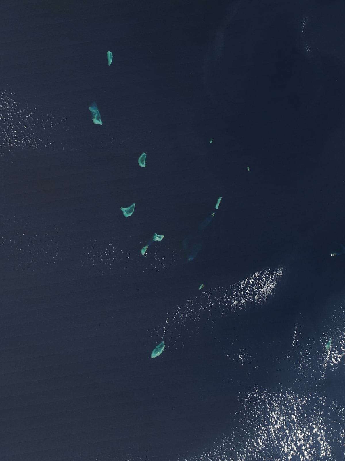 Satellite view of a dark blue sea dotted with a dozen small pale-green coral islands and reefs