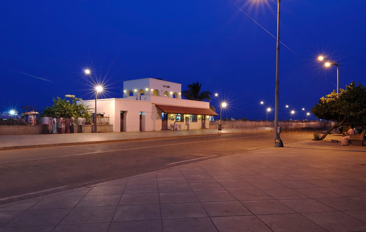 A wide paved seafront road at dusk with street lamps and a white building with arches