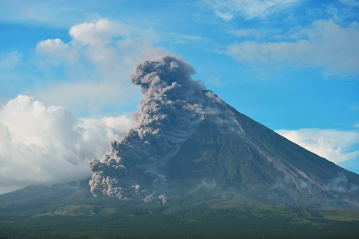 A tall, almost perfect cone-shaped mountain with a huge grey cloud of ash rolling down one side, under a blue sky