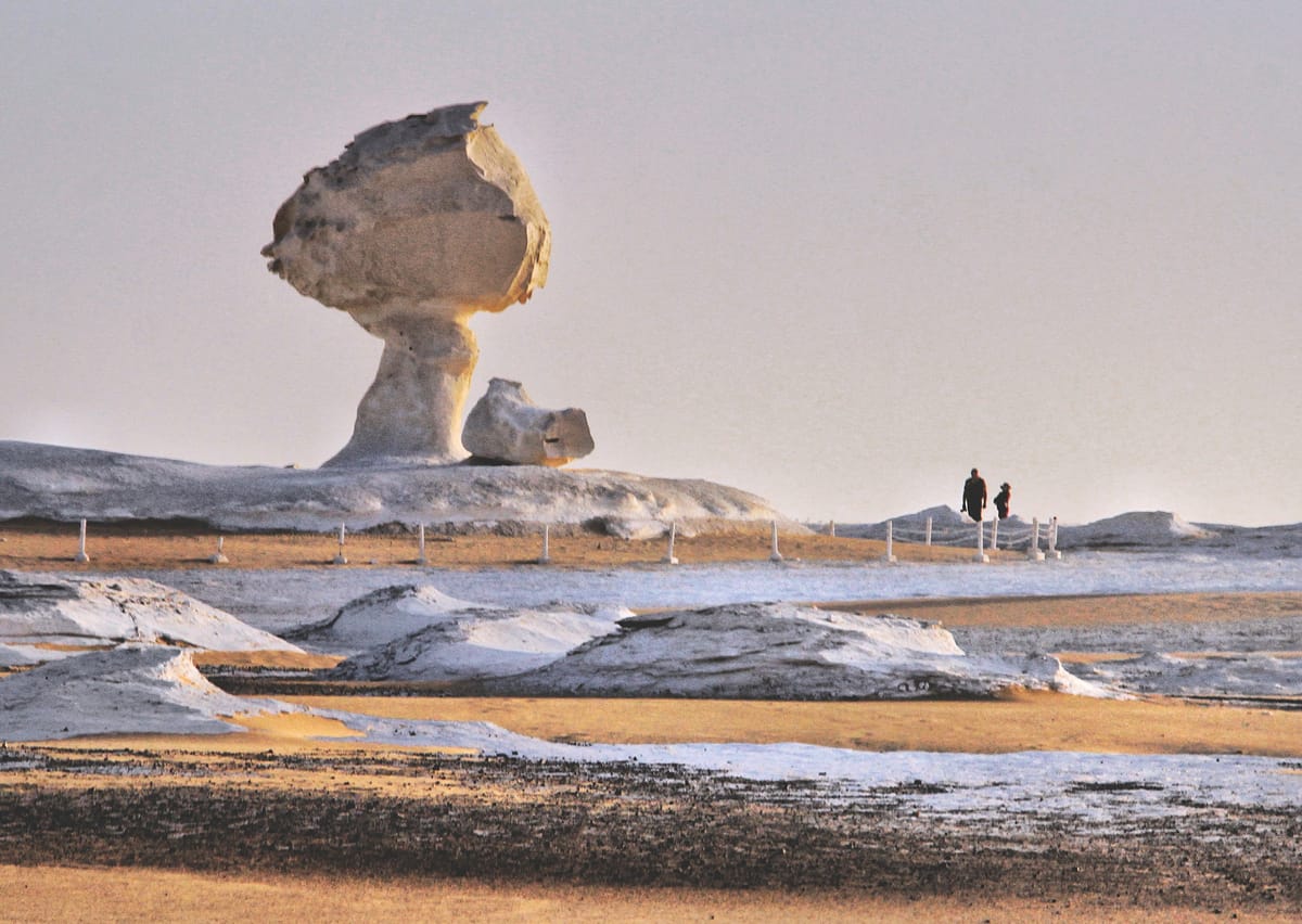 A large white rock shaped like a mushroom, with a thin stem and a wide top, standing in a flat desert with two people in the distance