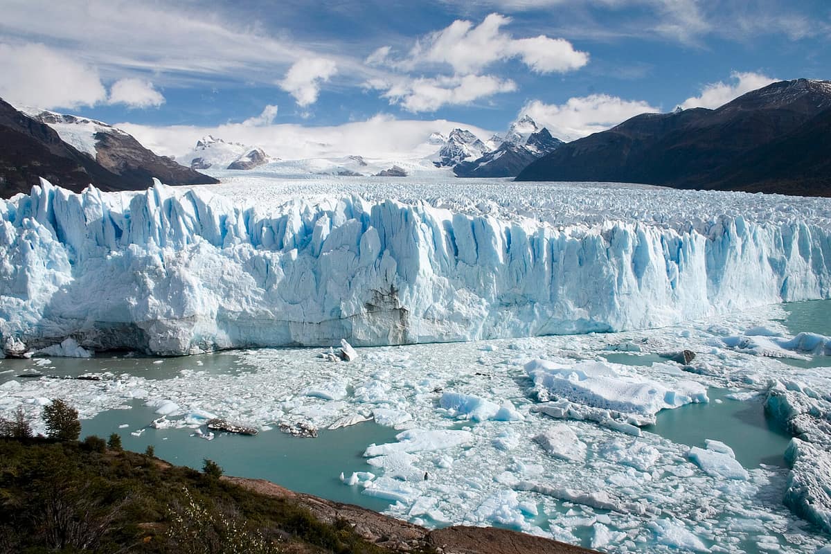 A wall of white and blue ice ending in a lake full of floating ice, with snowy mountains behind