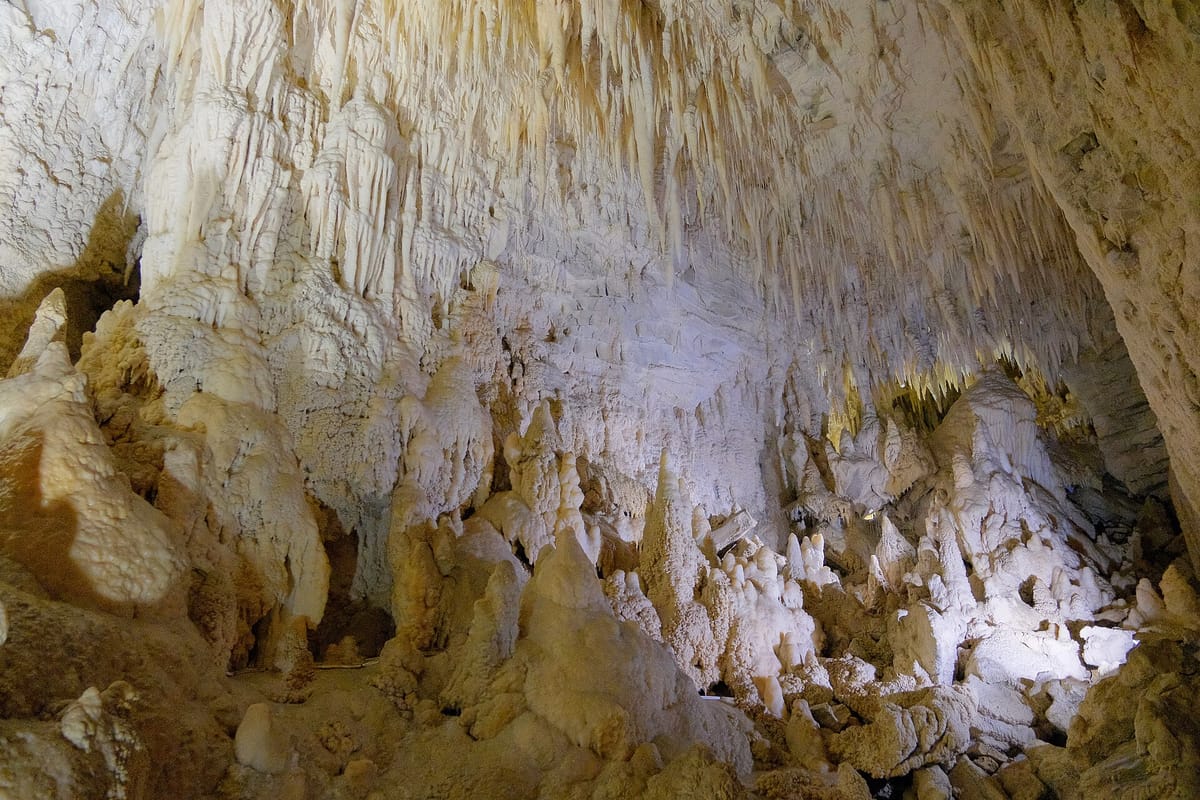 Inside a pale limestone cave, with many thin icicle-like stones hanging from the roof and lumpy columns rising from the floor