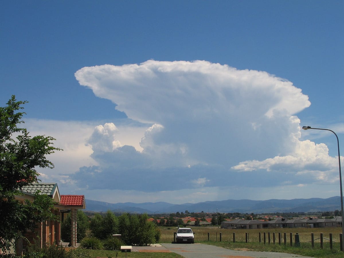 A huge white cloud tower rising high into a blue sky, flattened and spread out at the top like an anvil, above a row of houses and hills