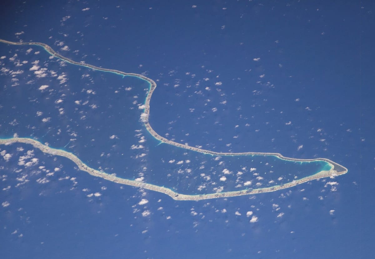 View from space of a long thin ring of pale land and pale-blue shallow water enclosing a large dark-blue lagoon in the open ocean, with small white clouds
