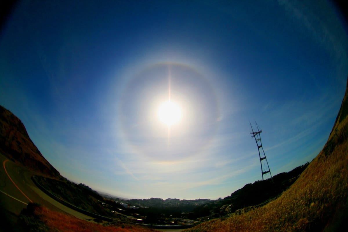 Wide-angle photo of the bright Sun in a blue sky, ringed by a faint circle of light, with a hillside and a tall tower below