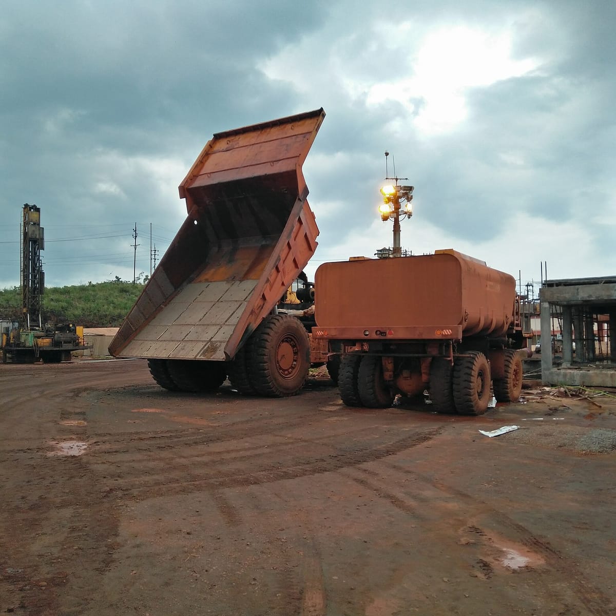 Two huge dumper trucks, one with its tipper raised, standing on muddy ground stained red by iron ore, under grey rain clouds