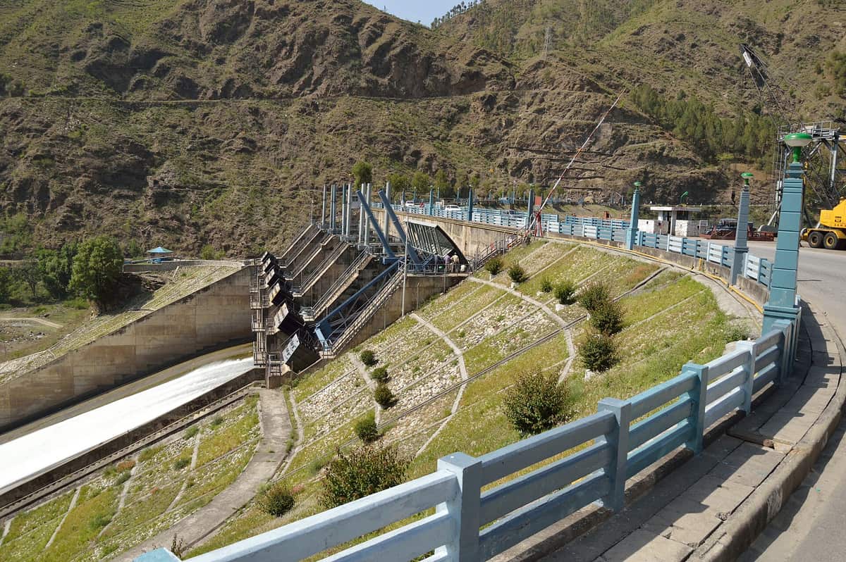 A concrete dam with tall steel floodgates across a river in a dry rocky valley, with water rushing down a spillway on the left and a road with blue railings on the right