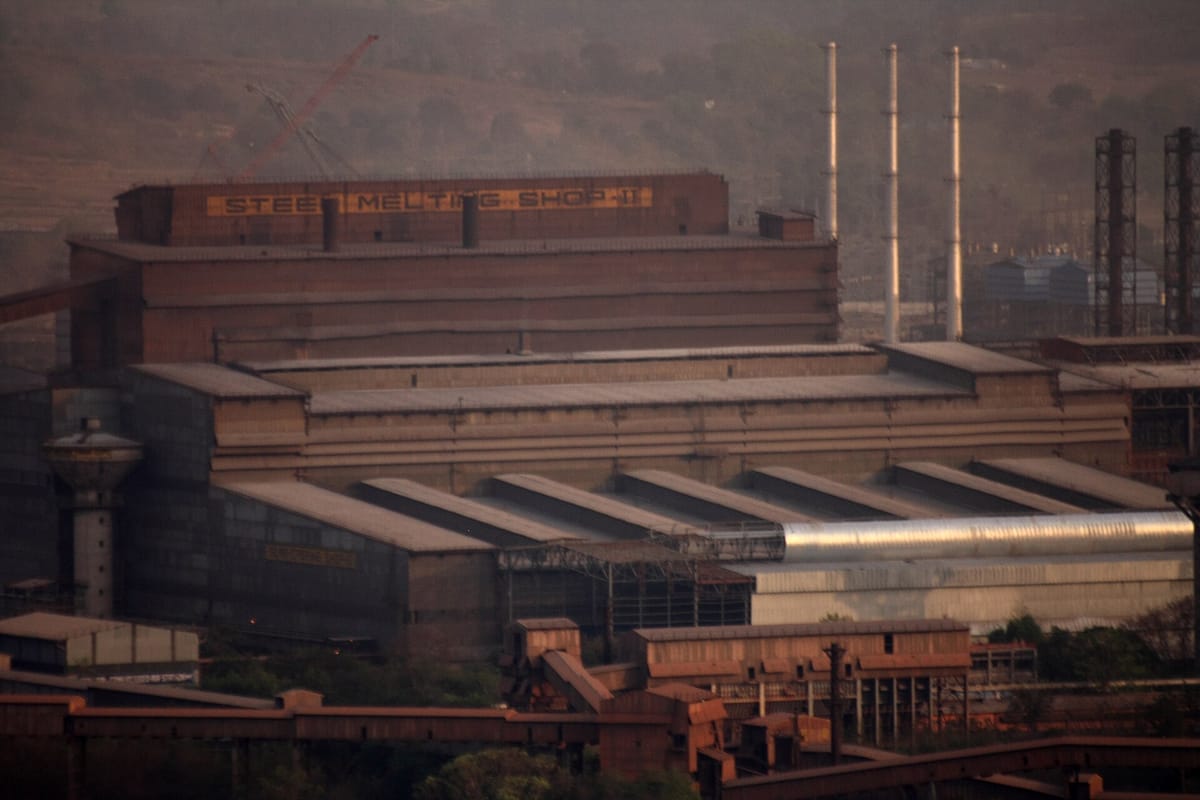 A huge brick-red factory building with 'Steel Melting Shop-II' painted along the top, tall thin chimneys and hills behind in evening haze