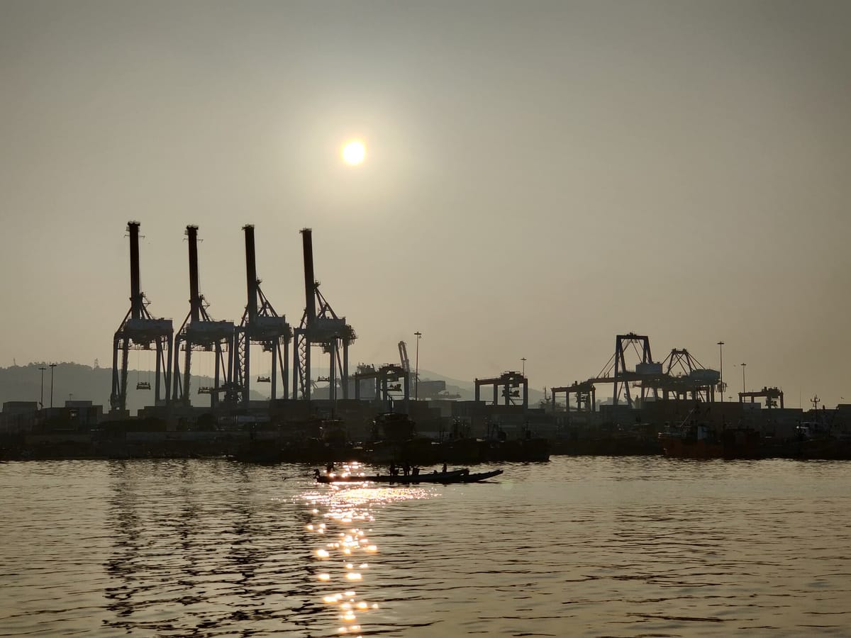 Tall container cranes and a dock seen in silhouette across calm water at sunrise, with a small fishing boat in front and hills behind