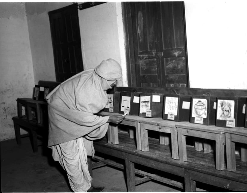 1952 ਵਿੱਚ ਇੱਕ ਵੋਟਰ ਬੈਲਟ ਬਕਸਿਆਂ ਦੀ ਕਤਾਰ ਅੱਗੇ ਖੜ੍ਹਾ, ਹਰ ਬਕਸੇ ਉੱਤੇ ਪਾਰਟੀ ਦਾ ਚੋਣ ਨਿਸ਼ਾਨ