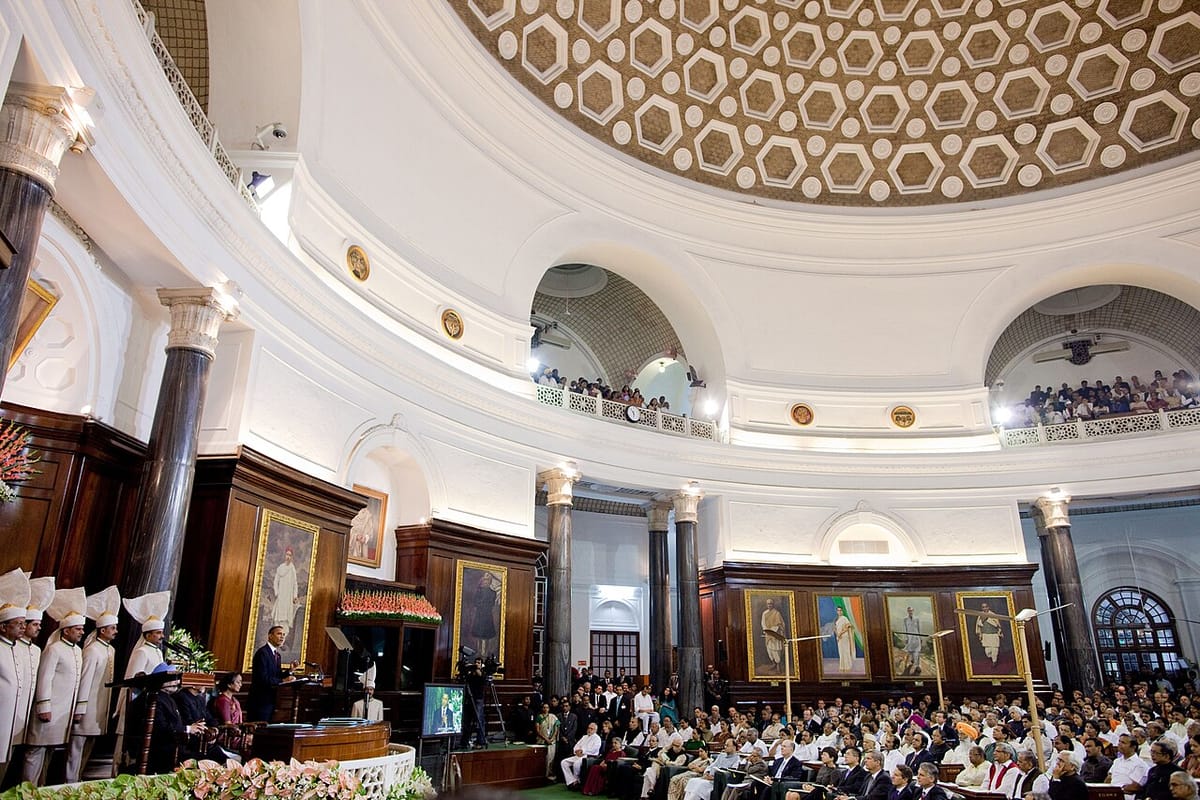 A large semicircular hall full of seated members listening to a speaker at a podium under a portrait