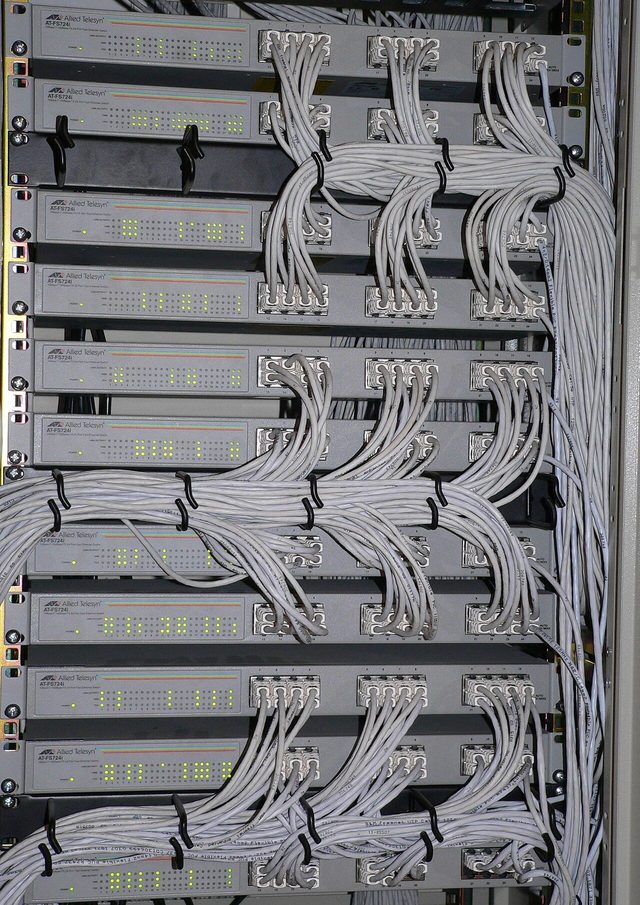 A tall metal rack holding network switches with many blue and grey cables plugged in