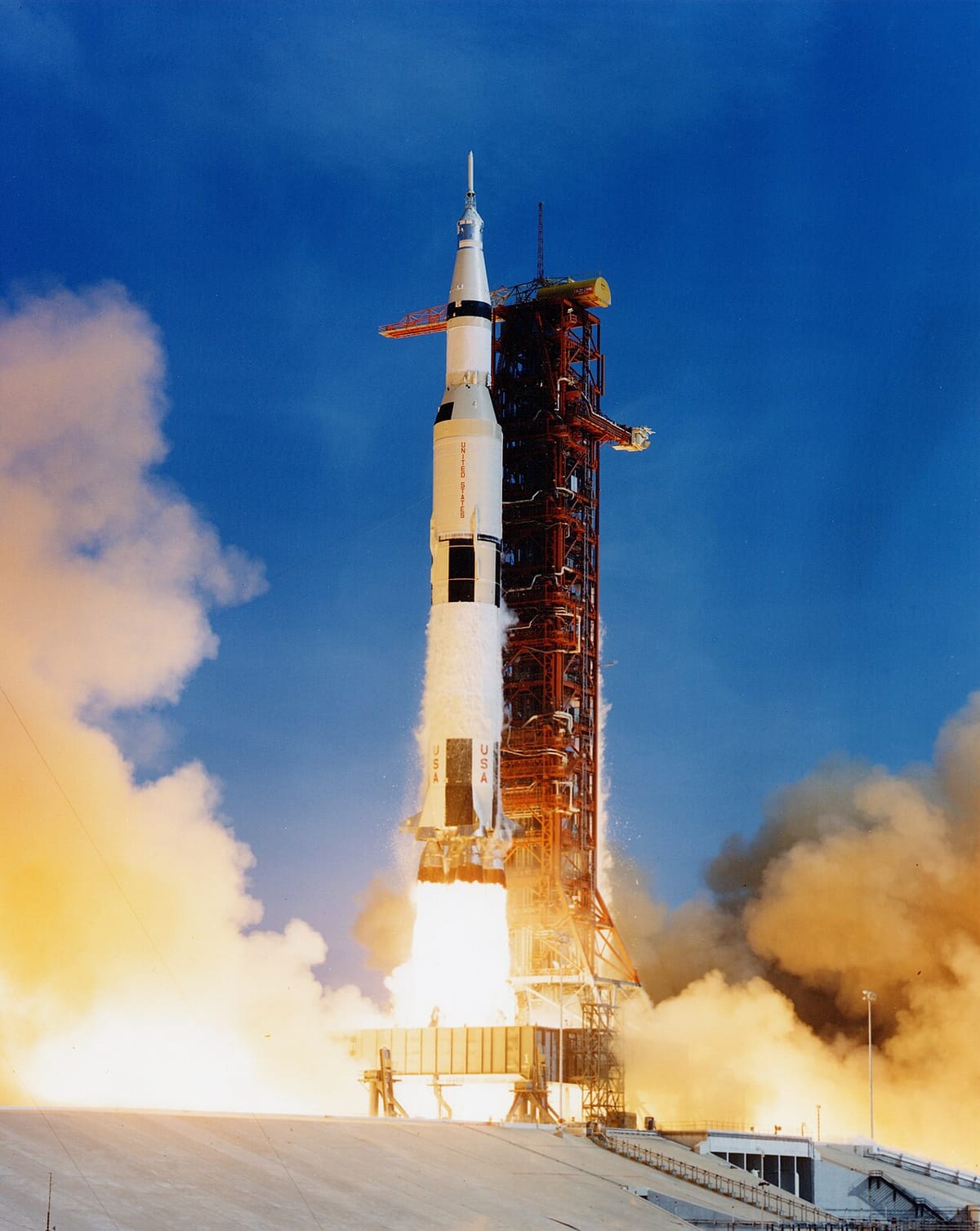 A tall white rocket lifting off its launch tower, with a huge burst of flame and smoke below it