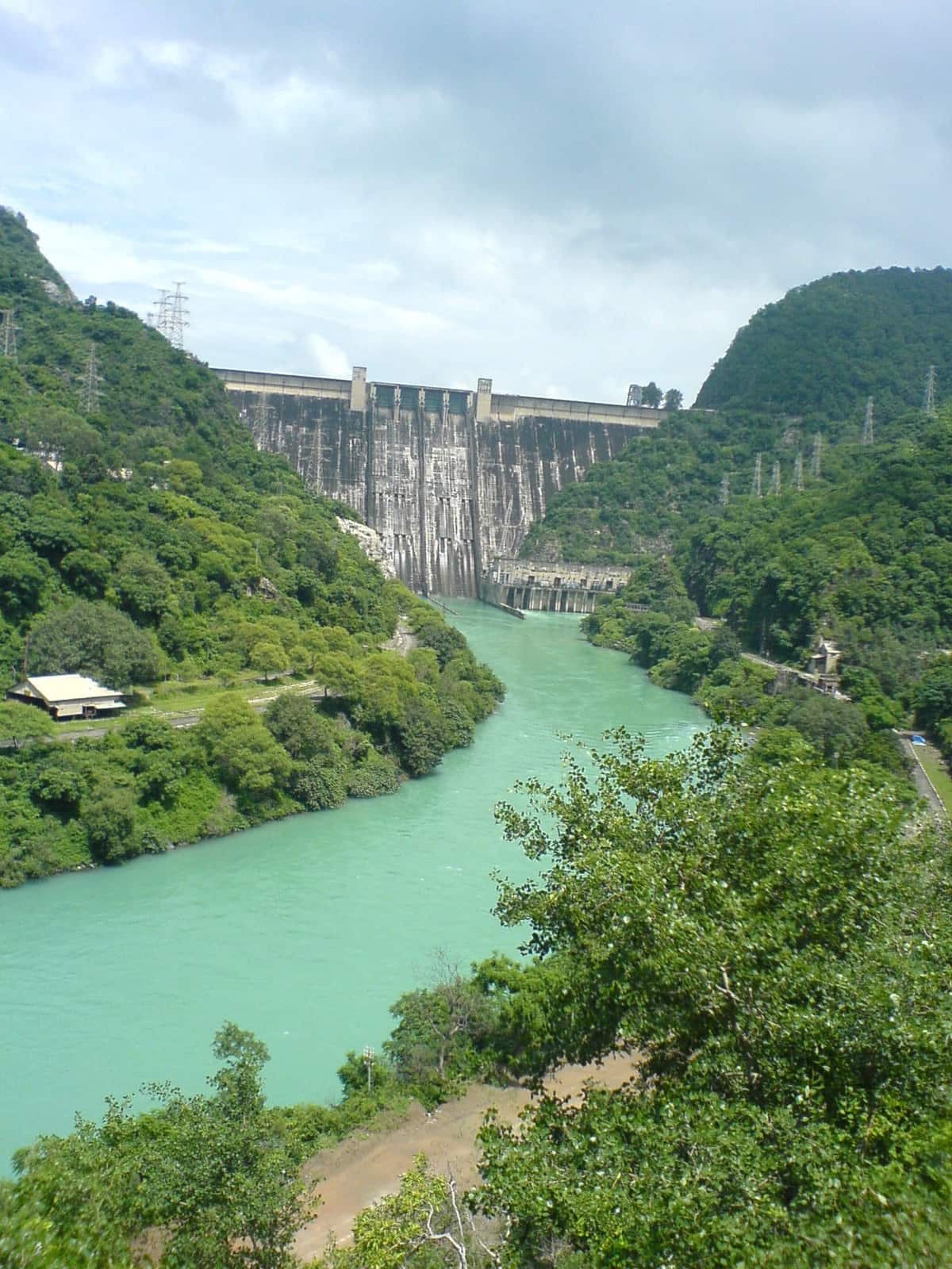 A tall concrete dam between green hills, with a blue-green river flowing away from it and power lines on the slopes