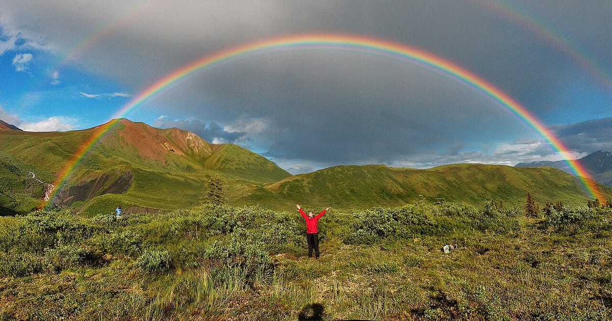 A bright full rainbow arching over green hills under a grey sky, with a fainter second rainbow outside it and a person with raised arms below