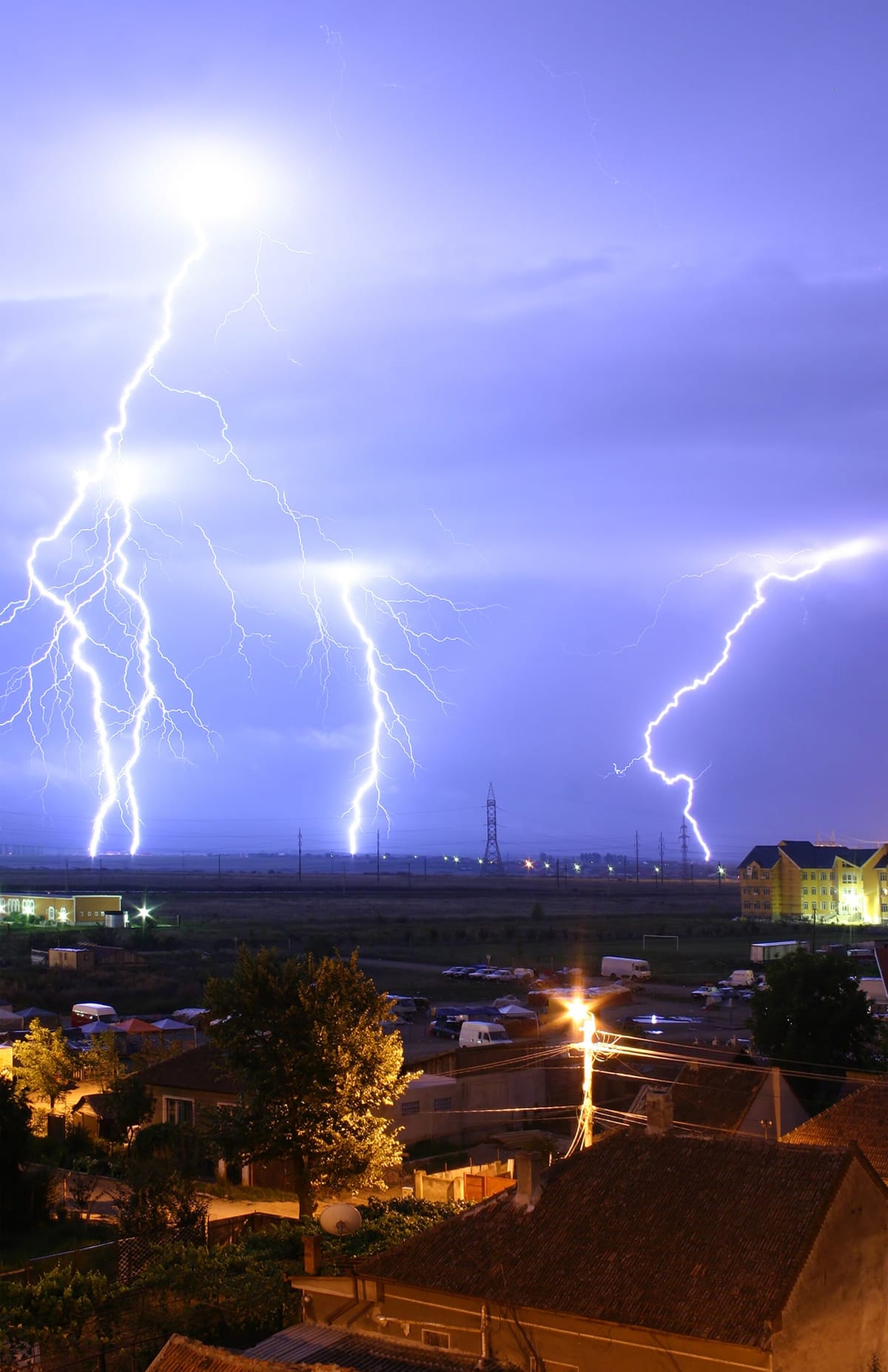 Several bright branching lightning bolts striking the ground at night beyond the roofs of a town