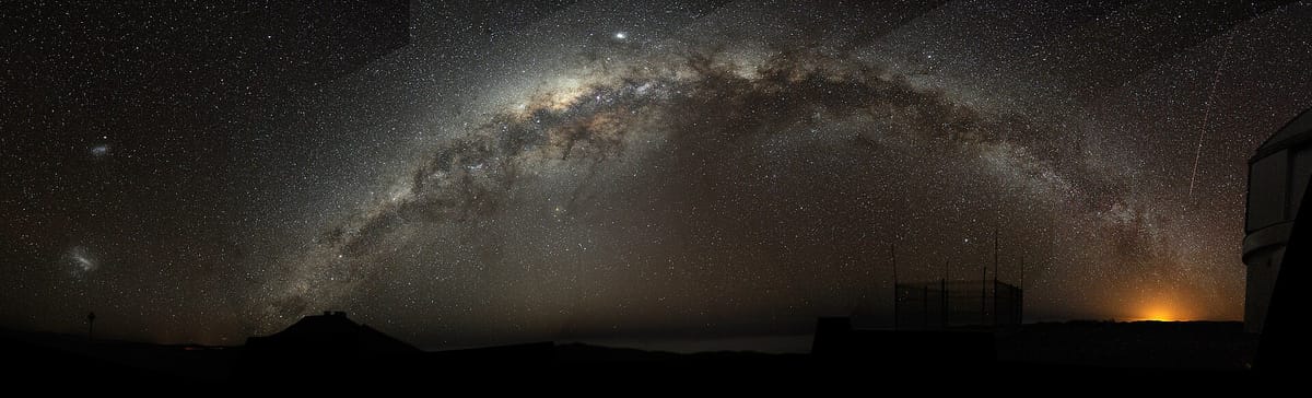A wide night-sky panorama with a bright band of countless stars and dark dust arching over dark hills and a telescope dome
