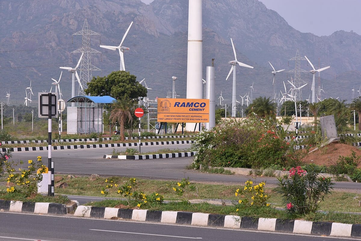 Many tall white three-bladed wind turbines in front of rocky hills, beside a road and an orange signboard
