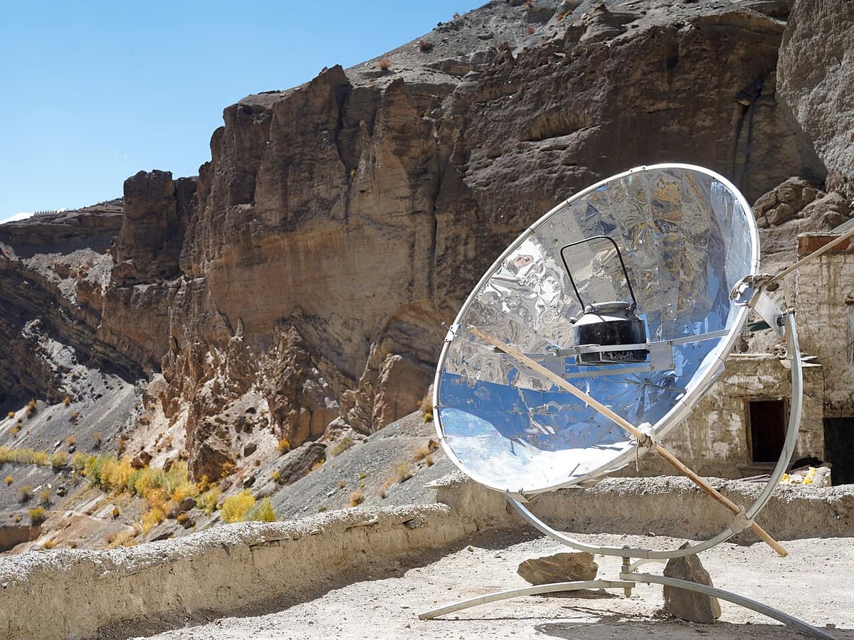A big round shiny dish mirror on a metal stand, with a black kettle held at its centre, on a rooftop below rocky cliffs