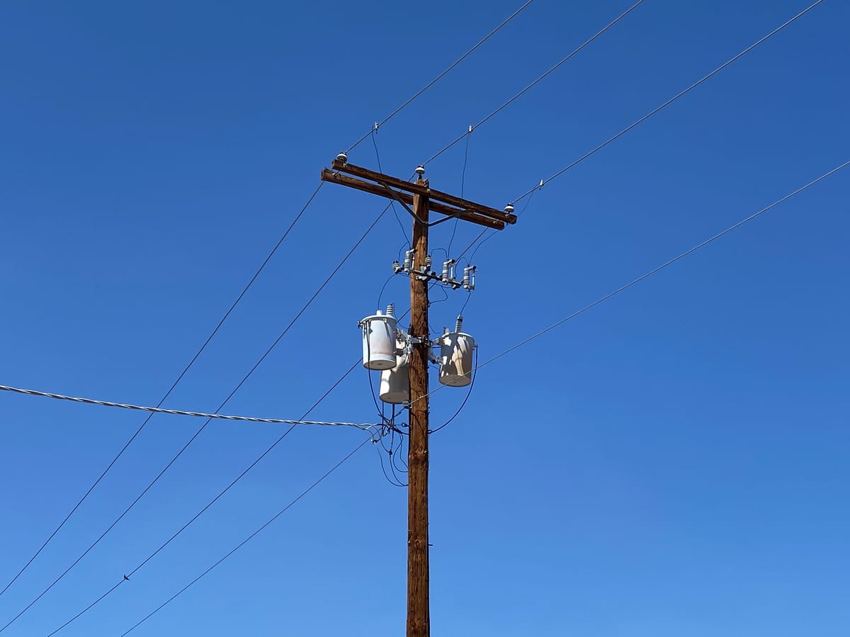 A wooden electric pole against a blue sky, carrying three grey drum-shaped transformers and many wires