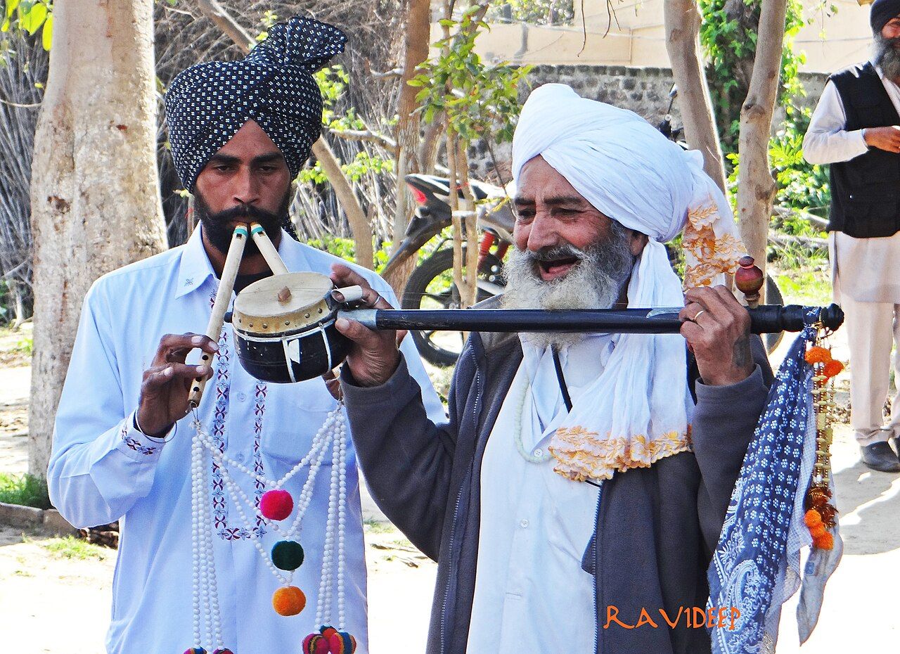 ਇੱਕ ਸਾਜ਼ਿੰਦਾ ਅਲਗੋਜ਼ੇ (ਲੱਕੜ ਦੀਆਂ ਦੋ ਬੰਸਰੀਆਂ) ਵਜਾ ਰਿਹਾ ਹੈ ਅਤੇ ਨਾਲ ਇੱਕ ਬਜ਼ੁਰਗ ਗਾਇਕ ਲੰਮੀ ਡੰਡੀ ਵਾਲਾ ਤੂੰਬਾ ਵਜਾ ਰਿਹਾ ਹੈ