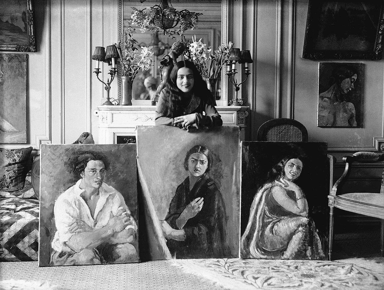Black-and-white photograph of Amrita Sher-Gil standing beside three of her paintings