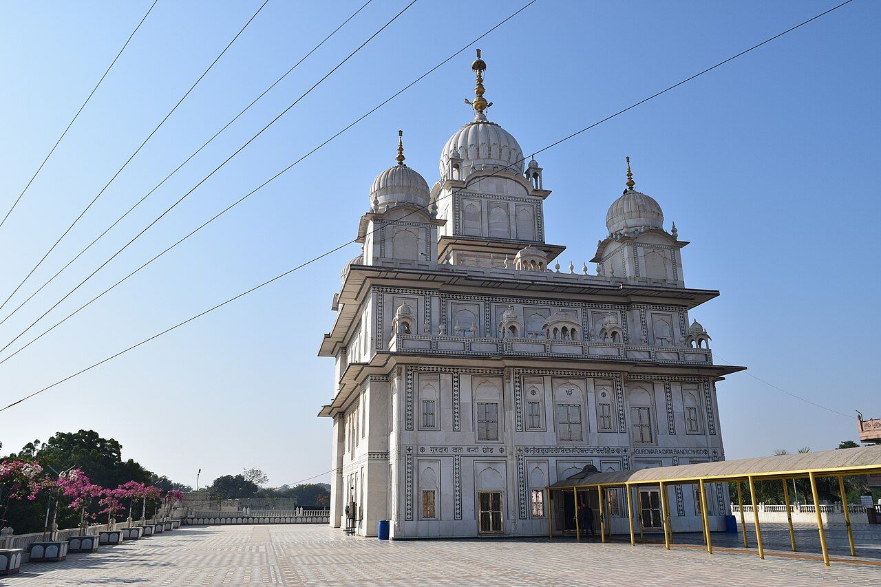 A tall white gurdwara with three domes on a stone terrace under a blue sky, at Gwalior Fort