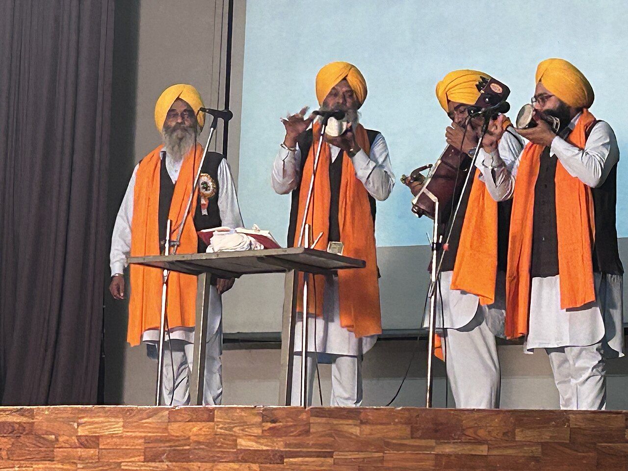 ਸਟੇਜ ਉੱਤੇ ਪੀਲੀਆਂ ਪੱਗਾਂ ਅਤੇ ਕੇਸਰੀ ਪਰਨਿਆਂ ਵਾਲੇ ਚਾਰ ਸਿੱਖ ਗਾਇਕ; ਦੋ ਦੇ ਹੱਥ ਛੋਟੀਆਂ ਢੱਡਾਂ ਅਤੇ ਇੱਕ ਸਾਰੰਗੀ ਵਜਾ ਰਿਹਾ ਹੈ