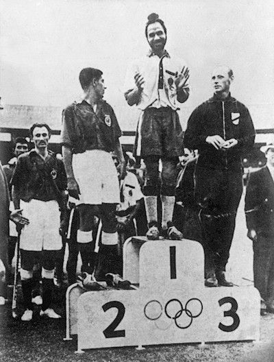 Black-and-white photograph of a hockey victory ceremony at the 1956 Olympics, with a Sikh player on the top step of the podium