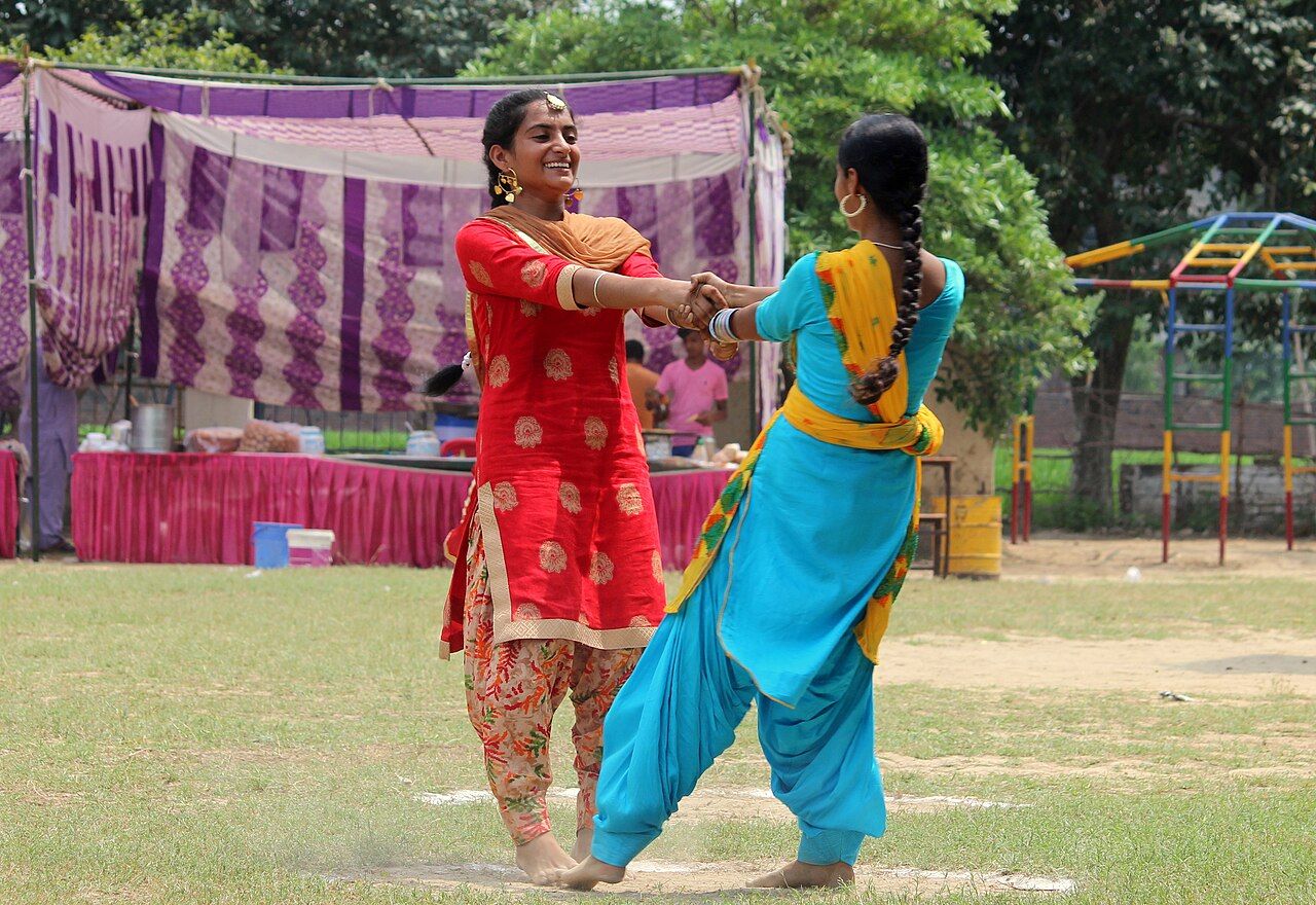 ਘਾਹ ਵਾਲੇ ਮੈਦਾਨ ਵਿੱਚ ਬਾਹਾਂ ਕਰਾਸ ਕਰਕੇ ਹੱਥ ਫੜ ਕੇ ਘੁੰਮਦੀਆਂ ਦੋ ਮੁਟਿਆਰਾਂ