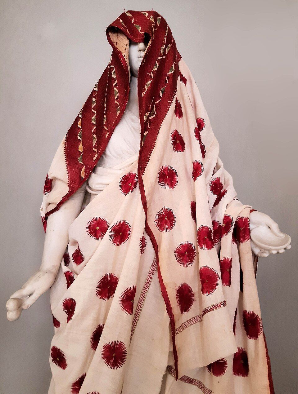 A cream khaddar phulkari shawl with red flower motifs and a red embroidered border, draped on a statue
