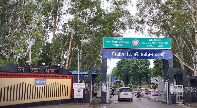 Entrance gate with a green sign reading Welcome Rail Coach Factory Kapurthala, on a tree-lined road