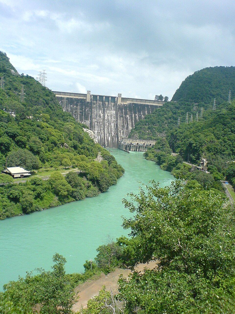 Bhakra Dam seen from downstream, with the green-blue Sutlej flowing out below it between wooded hills