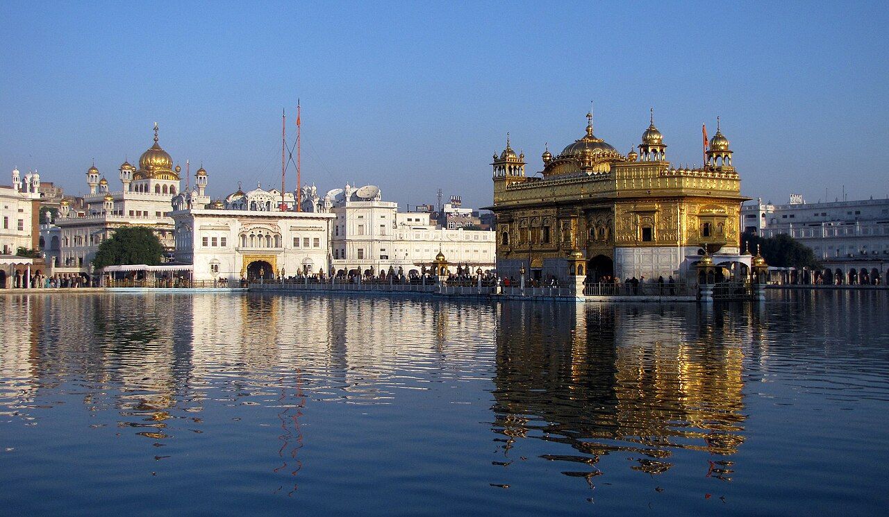 The gilded Harmandir Sahib in the middle of its sarovar, with white buildings around the tank, Amritsar