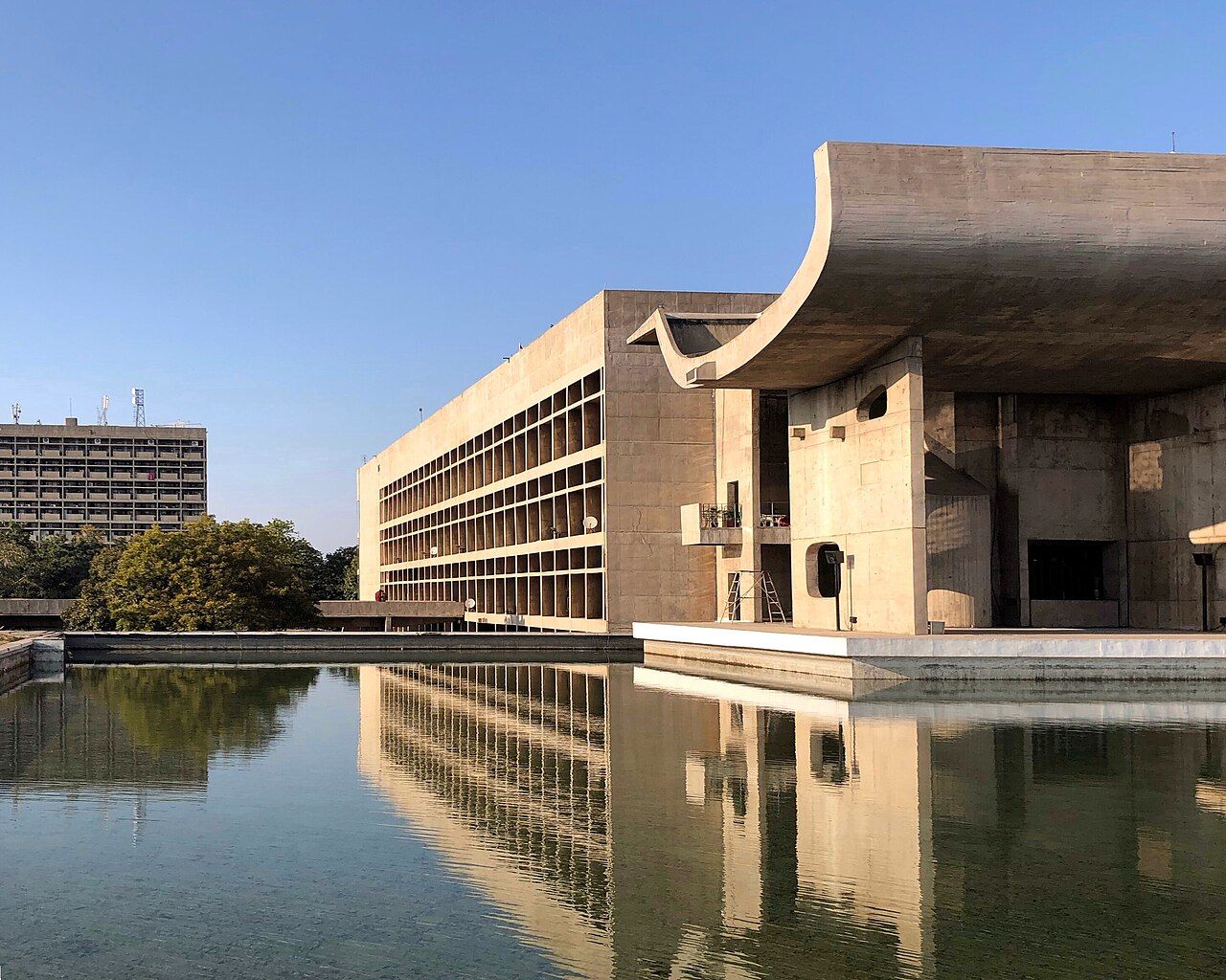 The Palace of Assembly in Chandigarh's Capitol Complex with its curved concrete roof, reflected in a pool, and the long Secretariat building behind