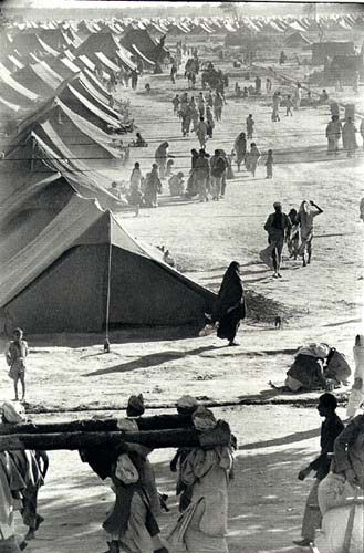 Black-and-white photograph of rows of tents in a refugee camp, with people carrying their belongings